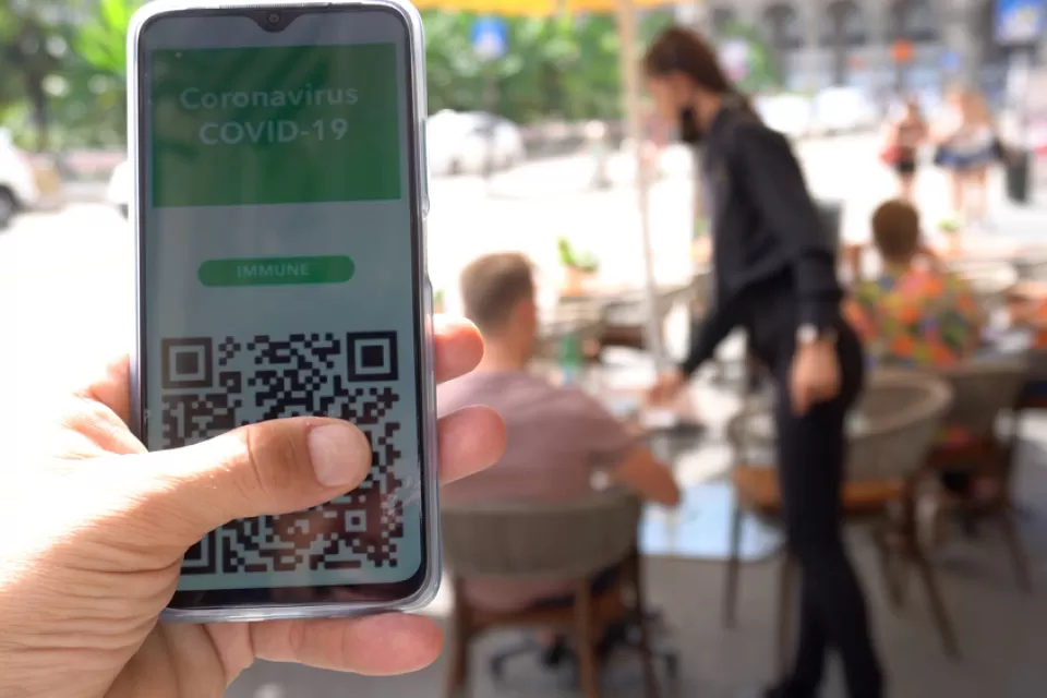 A digital vaccination record appears on a smartphone in front of a blurred background showing diners in an outdoor setting.